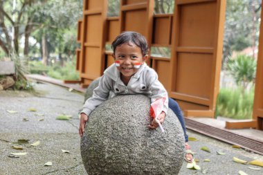 Bandung, Endonezya 29 Nisan 2025. Yüzü boyalı ve bayraklı neşeli çocuk parkta büyük bir taş kürede oynuyor..