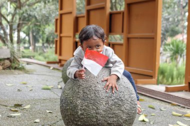 Bandung, Endonezya 29 Nisan 2025, Oyuncak çocuk taştan bir topun üzerinde yatarken kırmızı ve beyaz bayrak tutuyor..