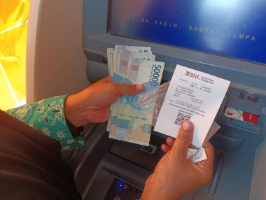 Bandung, Endonezya - 09 Mayıs 2025: Çekildikten sonra ATM nakit makbuzu olan ve Endonezya rupia banknotlarını ve dijital işlem fişini gösteren kişi