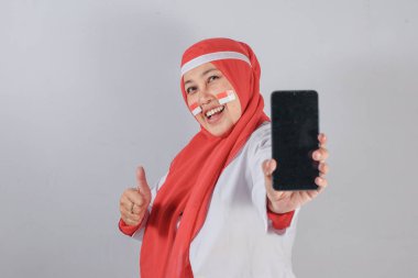 Endonezyalı, vatansever yüzlü bir kadın akıllı telefonuyla selfie çekerken neşeli bağımsızlık kutlamalarında gülümsüyor ve başparmağını kaldırıyor.