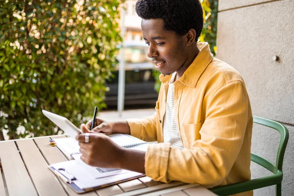 Side view of young African American man having a video call. He is talking from a bar and writing down the while studying online course
