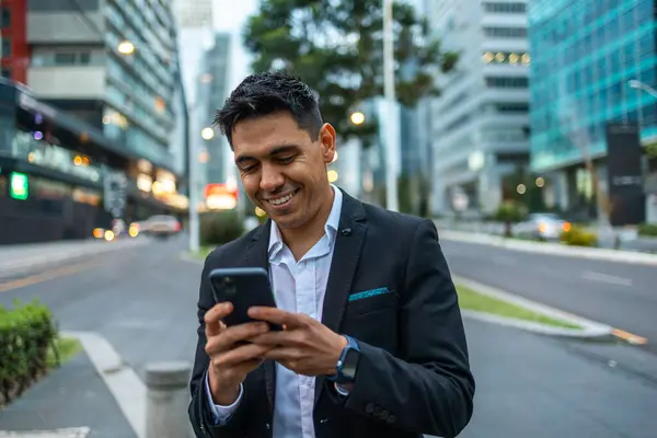 A portrait of a young Mexican smiling, happy, successful business man, executive walking outside down the street and using mobile phone