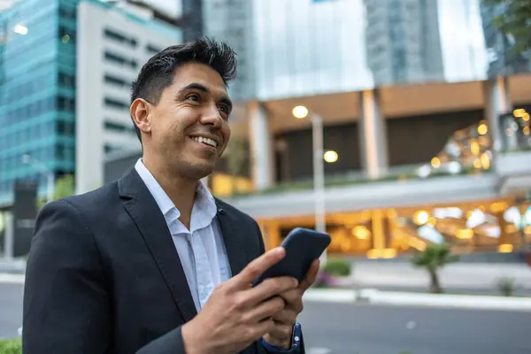 A portrait of a young Mexican smiling, happy, successful business man, executive walking outside down the street and using mobile phone