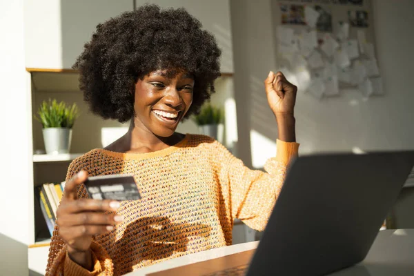 Excited young black woman using credit card and laptop for online shopping at home