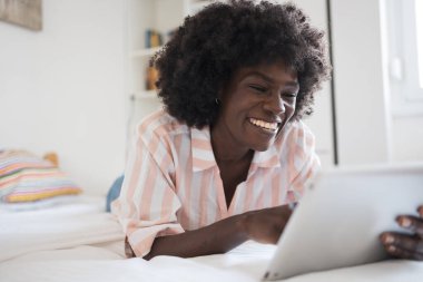 Evde yatarken dijital tablet kullanan afro saçlı neşeli siyah kadın. 