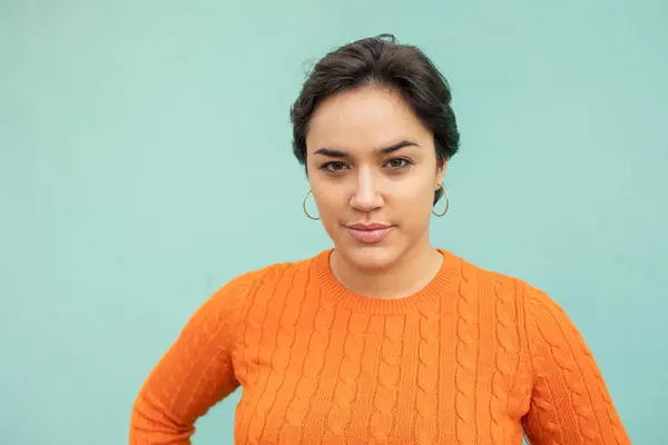 Portrait of confident young Latin woman in orange sweater against turquoise wall
