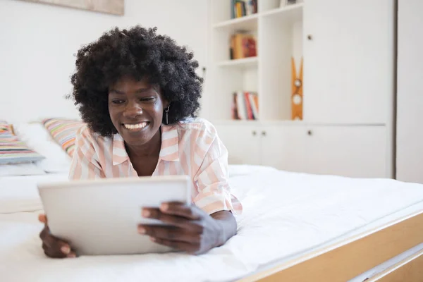 Happy young black woman with afro hairstyle browsing on digital tablet while lying in bed at home