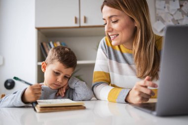 Gülümseyen, sarı saçlı serbest çalışan, dizüstü bilgisayarda çalışan ve oğlunun evde kitap yazmasına bakan genç bir anne.