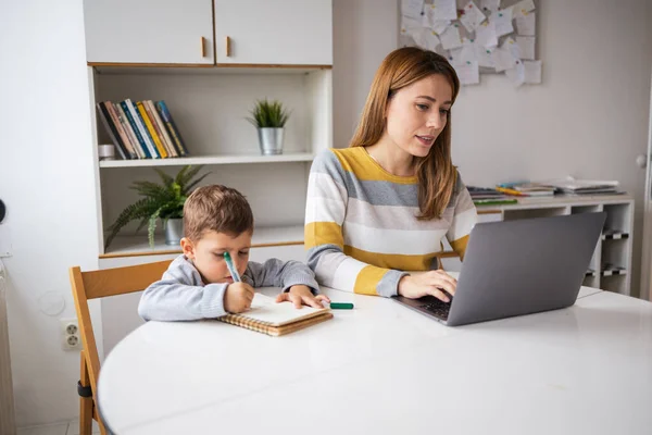 Young stay at home mother in casual clothes using laptop sitting beside son doing homework at desk at home