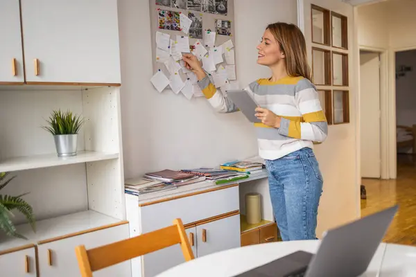 Smiling young female freelancer in casual clothes holding digital table while analyzing paper memos on notice board at home