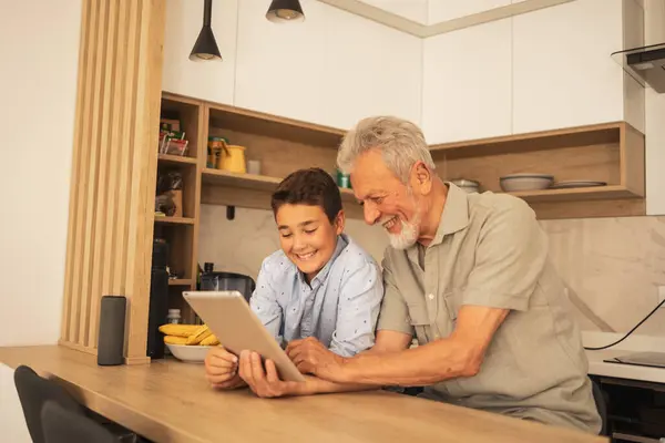 Smiling old grandpa with boy in casual clothes using digital tablet together at wooden table in kitchen at home during weekend