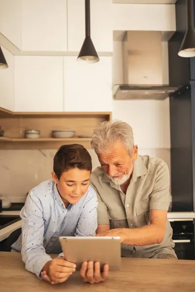 Happy adorable boy teaching grandfather to use digital tablet at wooden table in kitchen at home during weekend