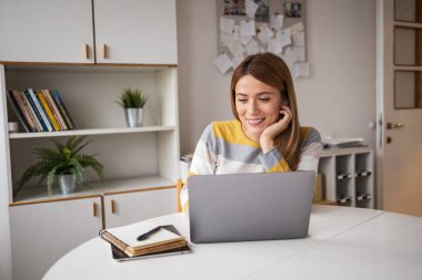 Günlük giysiler içinde gülümseyen genç bir kadın ofis masasında dizüstü bilgisayar kullanırken çenesi üzerinde oturuyor.