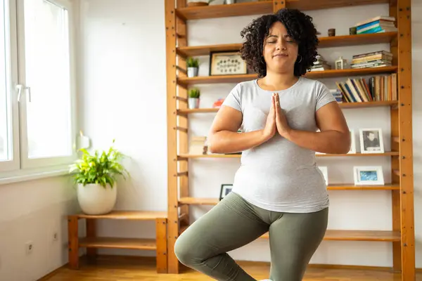 Relaxed young Latin woman in sportswear smiling while practicing tree pose in yoga room at home
