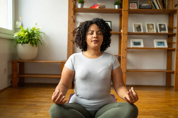 Relaxed young Latin woman practicing lotus pose while sitting on floor in yoga room at home