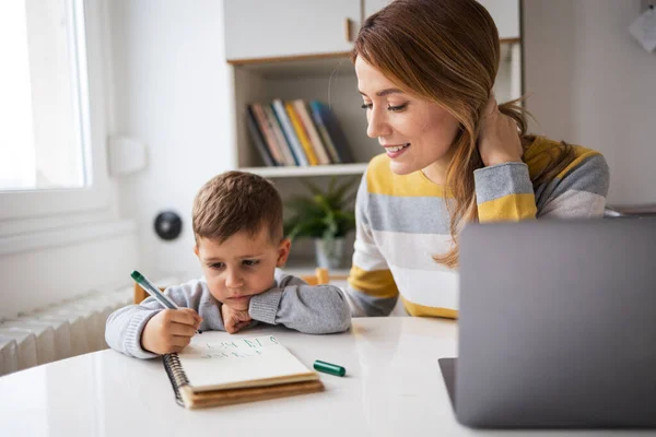 Smiling young working mother with laptop assisting son with homework sitting at desk at home