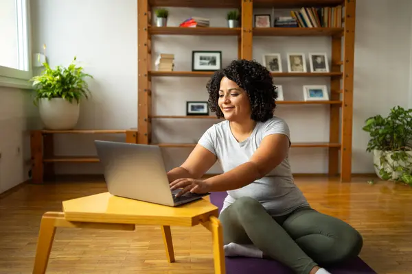 Full body of happy young Latin woman using laptop while sitting on yoga mat at home