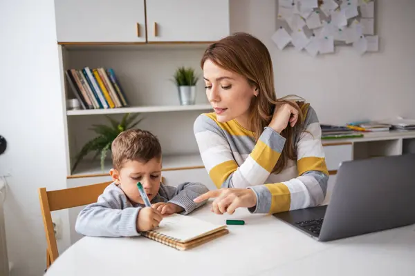 Young blond working mother in casual clothes with laptop assisting son with homework at desk at home