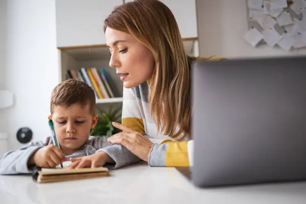 Focused young blond working mother with laptop assisting son doing homework at desk at home
