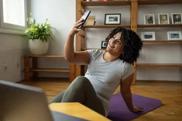 Happy young Latin woman in activewear taking selfie through cell phone while sitting on yoga mat at home