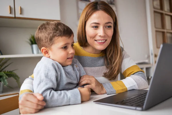 Smiling young woman and son in casual clothes looking at laptop screen while sitting at desk at home