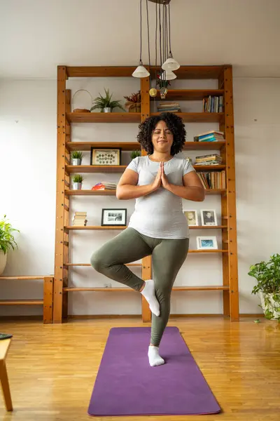 Full body of relaxed young Latin woman in sportswear practicing tree pose on yoga mat at home