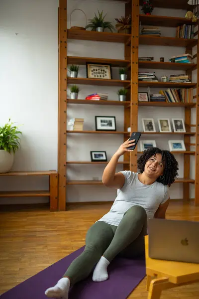 Full body of happy young Latin woman taking selfie through cell phone while sitting on yoga mat at home