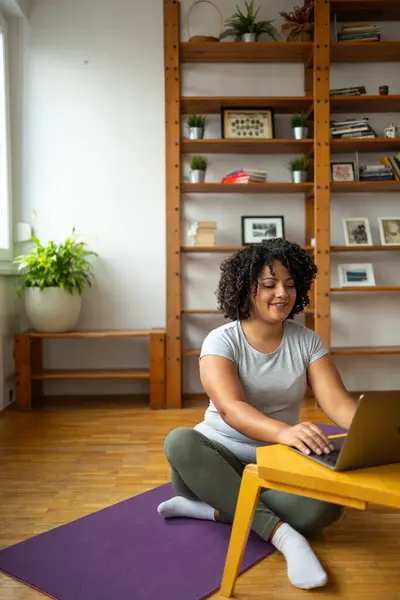 Full body of happy young Latin woman using laptop while sitting with legs crossed on yoga mat at home