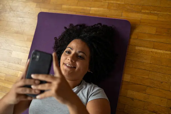 就在上面 一个快乐的拉美女人躺在瑜伽垫上用智能手机拍到的照片 — 图库照片