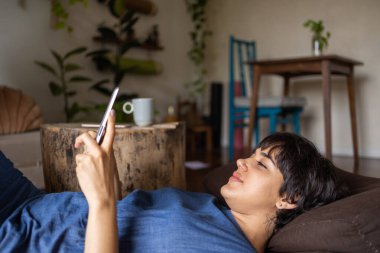 Telefonunda kısa saçlı mesajlaşan rahat, genç bir Latin kız. Yeşil bitkilerle dolu bir odada gevşemiş mavi bir elbise giyiyor. Yüksek kalite fotoğraf