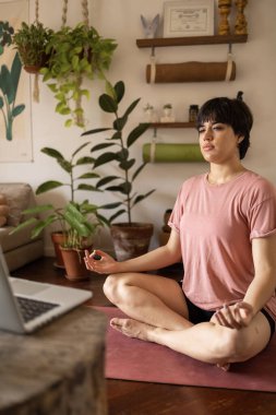 Laptopa bakarken kısa saçlı meditasyon yapan Latin bir kadının dikey hali. Pembe gömlek giyiyor ve yeşil bitkilerle dolu bir odada oturuyor..