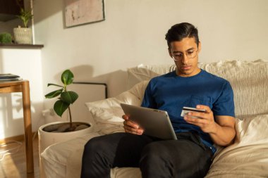 Gözlüklü, mavi gömlekli, tablet kullanarak koltukta oturan ve kredi kartına bakan çekici İspanyol bir adam. Yüksek kalite fotoğraf