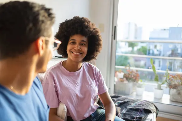 stock image Cheerful latin couple talking in the living room with a window in the background. They are siting on the sofa in apartment with lot of natural light. Girl has afro hair and big smile.