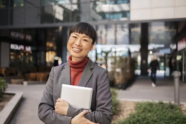 stock image Smiling Japanese businesswoman holding a tablet outdoors in a modern office complex, exuding confidence and professionalism.