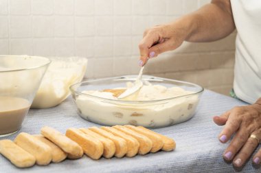 Bir el, ıslak kadın parmaklarını cam bir tabakta kremalı mascarpone ile kaplıyor ve İtalyan tatlısı Tiramisu 'nun yapımında son adımı atıyor..