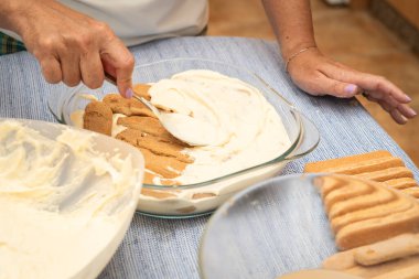 Bir kadın parmaklarının üzerine kremalı mascarpone kepçeleyen biri, geleneksel tiramisu tatlısını cam bir tabakta hazırlıyor, mutfak tezgahında hazırlanan malzemelerle birlikte.