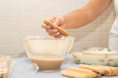 Bir hanımefendinin kahveye bandırdığı bir el resmi, mutfakta klasik bir tiramisu tatlısı hazırlıyor.