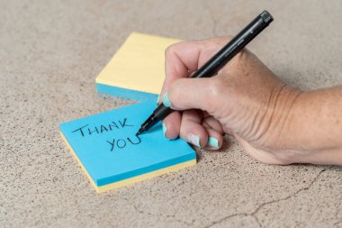 Siyah kalemle mavi yapışkan bir nota teşekkür yazan birinin yakın çekimi. Kişilerin elleri ve yapışkan notlar desenli bir yüzeyde görülebilir..