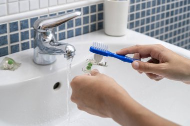 Diş fırçasını musluktan akan suyun altında diş fırçasıyla temizleyerek ağız hijyeni ve bakım masraflarını vurgulayan ellerin yakın çekimi