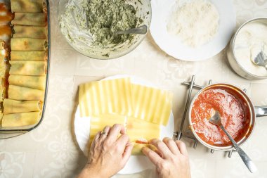 Bir insan, domates sosu, peynir ve beşamel gibi malzemelerle çevrili ev yapımı cannelloni hazırlarken makarna tabakalarına ricotta ve ıspanak karışımı sürer.