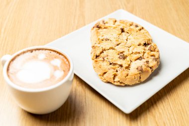 İki çikolatalı kurabiye, kapuçino bardağının yanında, içinde latte resmi olan, ahşap bir masaya yerleştirilmiş, samimi ve davetkar bir atmosfer yaratıyor..