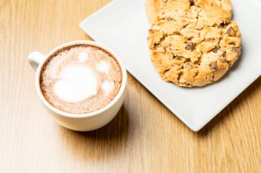 Kahve fincanı, latte sanatıyla süslenmiş, ahşap bir masada taze pişmiş çikolatalı kurabiyelerin yanında oturur, samimi ve davetkar bir sahne yaratır.