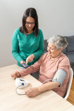 Kızı, yaşlı annesinin kan basıncını ölçmek için bir kan basıncı monitörü ve mobil bir uygulama kullanarak annesine uygulamayı ve kan basıncı monitörünü nasıl kullanacağını öğretiyor..