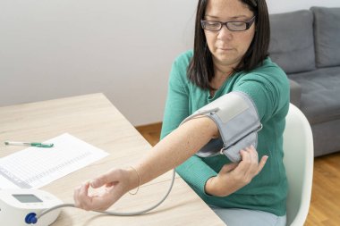 Kolundaki kan basıncını ayarlayan kadın, evde dijital gerilim ölçer ile ölçüm yapmaya hazırlanıyor.