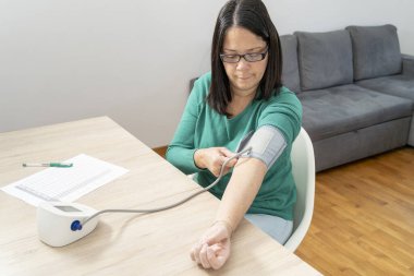 Kolundaki kan basıncını ayarlayan kadın, evde dijital gerilim ölçer ile ölçüm yapmaya hazırlanıyor.