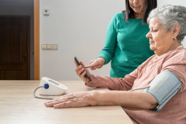 Hemşire yaşlı bir kadına tansiyonunu ölçmesi için yardım ediyor ve akıllı telefondan sonuçları kontrol ediyor.
