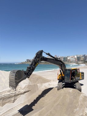 Kazıcı kumsalda kum taşıyor, arka planda bir şehir var.