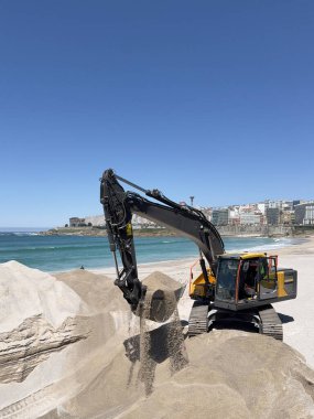 Güneşli bir günde, arka planda deniz ve şehir olan bir kumsalda kum taşıyan kazıcı.