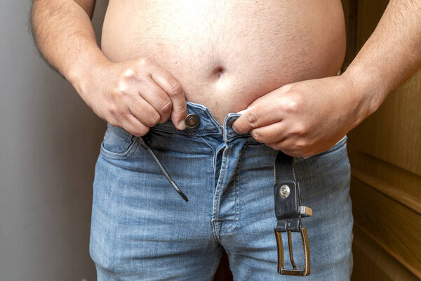 Мужчина с избыточным весом изо всех сил пытается закрыть свои узкие джинсы, представляя собой увеличение веса и проблемы с диетой