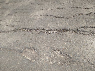 Çatlamış ve yıpranmış asfalt yol yüzeyinde görünür çukurlar ve küçük taşlar var..